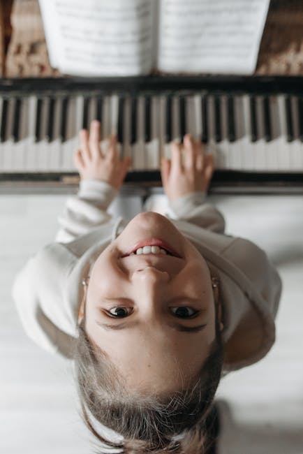 A joyful young girl in a hoodie smiles up at the camera while playing the piano.